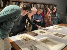 A large wood table had several matted prints laying on it. The prints are etchings and have been made in black ink. Standing over the table is a group of six people viewing the artworks. One of the points to a particular artwork while others look.