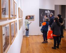 Gallery with white walls and wood floor featuring photographs framed along the wall behind glass. A tour guide speaks about the images to a groups of people dressed in winter clothing.