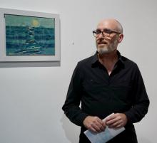 Wes Sherman wears a black collar shirt and stylish glasses. He has a bear and a shaved head, his hands hold a folded pamphlet. Sherman stands in front of a white wall that features a framed landscape painting.