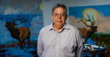 Artist Norman Akers stands in front of two paintings wearing a light blue collar shirt, glasses, and his hair tied back. His arms are behind his back. The two paintings in the background features elks amidst seas of garbage and devastated landscapes. The elks are crying out in what appears to be anguish.
