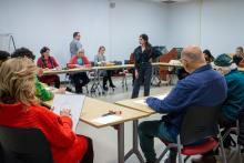 People sit at wood desks in a classroom while they create charcoal sketches in paper sketchbooks. The desks surround a model posing in all black.