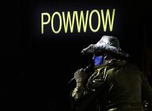 Photograph with black background and yellow text reading "Powwow". In the foreground is a presenter wearing a gold shirt, gold glove, and reflective cowboy hat with a blue mask or blue face makeup on. In the presenter's hand is a microphone.