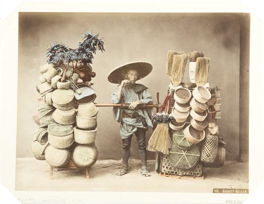 Handcolored studio photo of a young male Japanese vendor between two tall displays of baskets and brooms