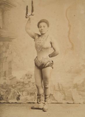 Sepia photo of a petite Black woman wearing a decorated leotard, standing and holding onto a trapeze with her right arm