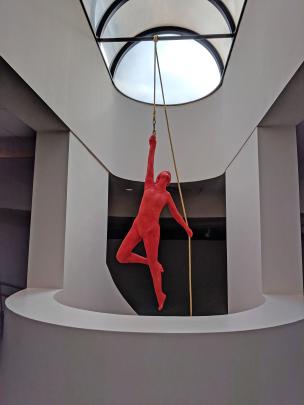 Life-sized sculpture of an acrobat painted red, hanging from ceiling by a yellow rope near a skylight 