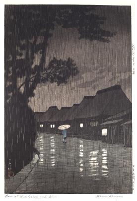 A lone figure walks through a Japanese village on a rainy night. Lights from windows glisten on the wet walkway. 