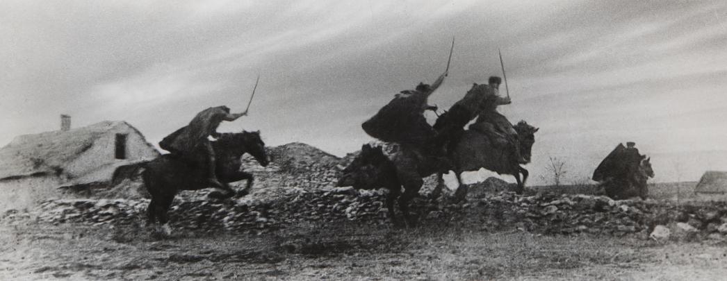Photo of four figures carrying swords riding horses jumping over a stone wall