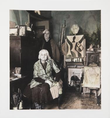 Photo of a studio filled with various art; a woman stands behind an older woman in a chair
