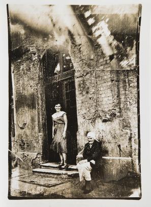 Black and white photo of a young woman in a dress standing in a doorway while an older woman sits to the side