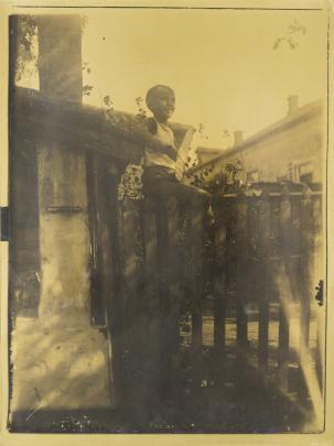 Yellow-tinted photograph of smiling boy standing on a fence in front of a house