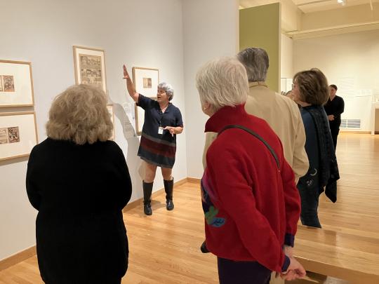 Two people watch as a tour guide points at artwork.