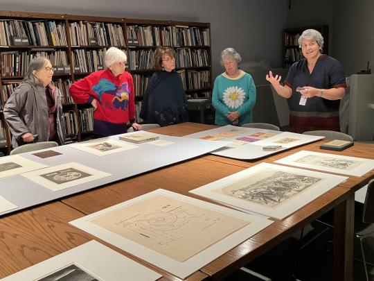 People stand around a table covered in artwork while a guide talks.