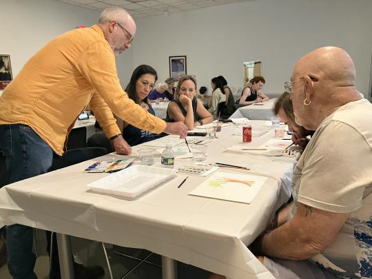 Artist Wes Sherman, wearing a bright yellow long sleeve shirt, leans on a table covered in painting supplies. Sitting at the table are various people that listen to Wes as he demonstrates painting technique.