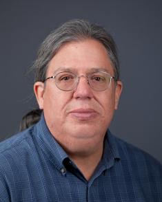 Headshot of Norman Akers in front of a blue and gray backdrop. He wears a blue collar shirt and glasses. His hair is tied back and his expression has a slight smile with serious eyes.