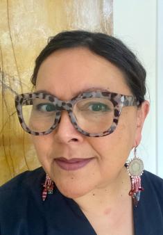 Selfie of Sonya Kelliher-Combs, a female presenting person wearing a navy v neck top, tortoise shell patterned large frame glasses, and decorative purple earrings. Her hair is black and pull back in a bun or ponytail. She is lightly smiling and her eyes look toward the camera.