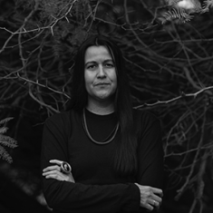 Natalie Diaz wears all black with her arms crossed in front of dead branches. She has a serious expression emphasized by her minimalistic jewelry and straight black hair.